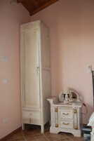 Una habitación con un armario alto de color blanco, una mesita de noche pequeña con una lámpara y un suelo de baldosas.