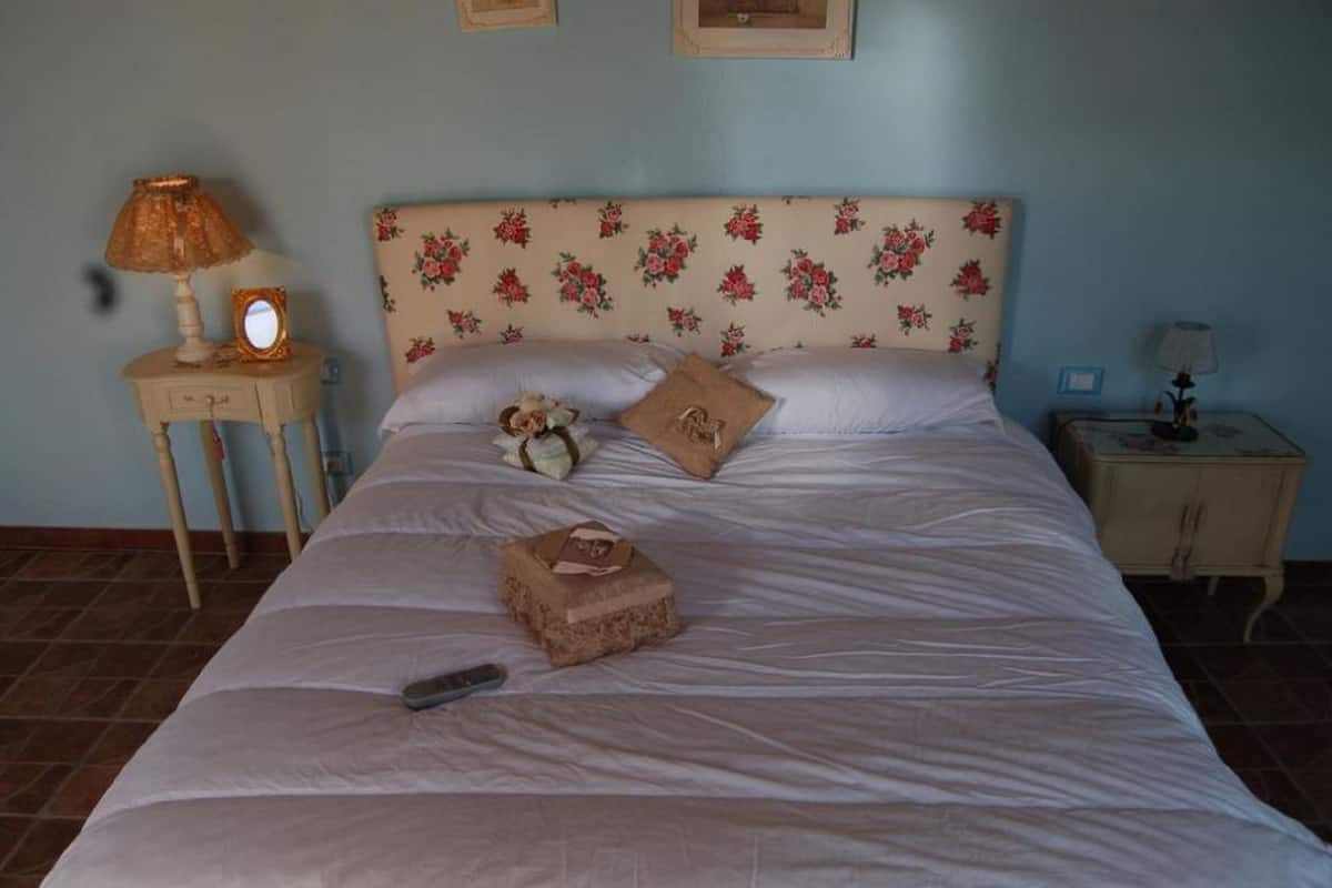 A bedroom with a floral headboard, two bedside tables with lamps, a bed with a remote control, and a small box on the bed.