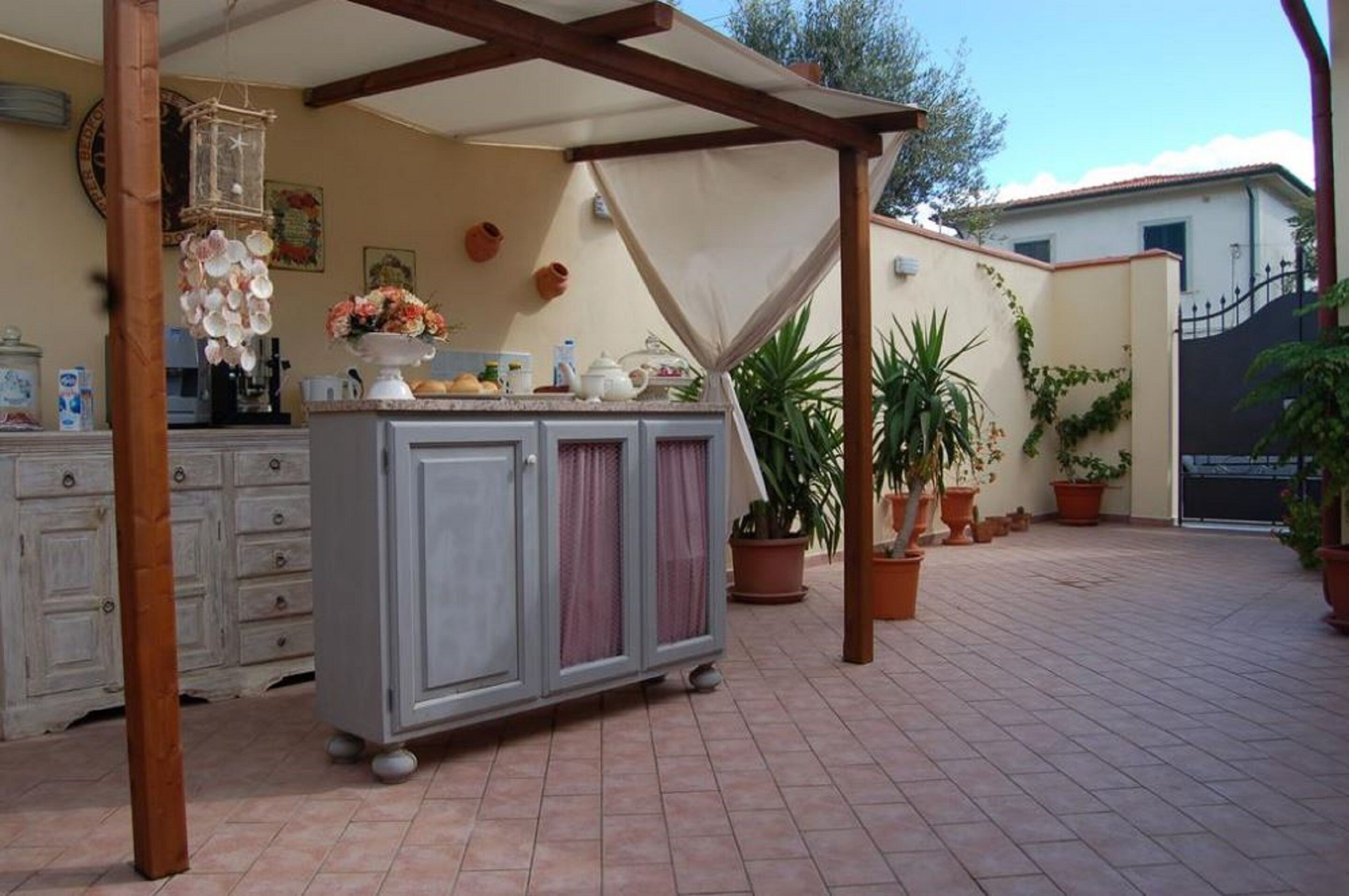 A covered outdoor area with a tiled floor, a wooden pergola, a cabinet with kitchen items, and potted plants.
