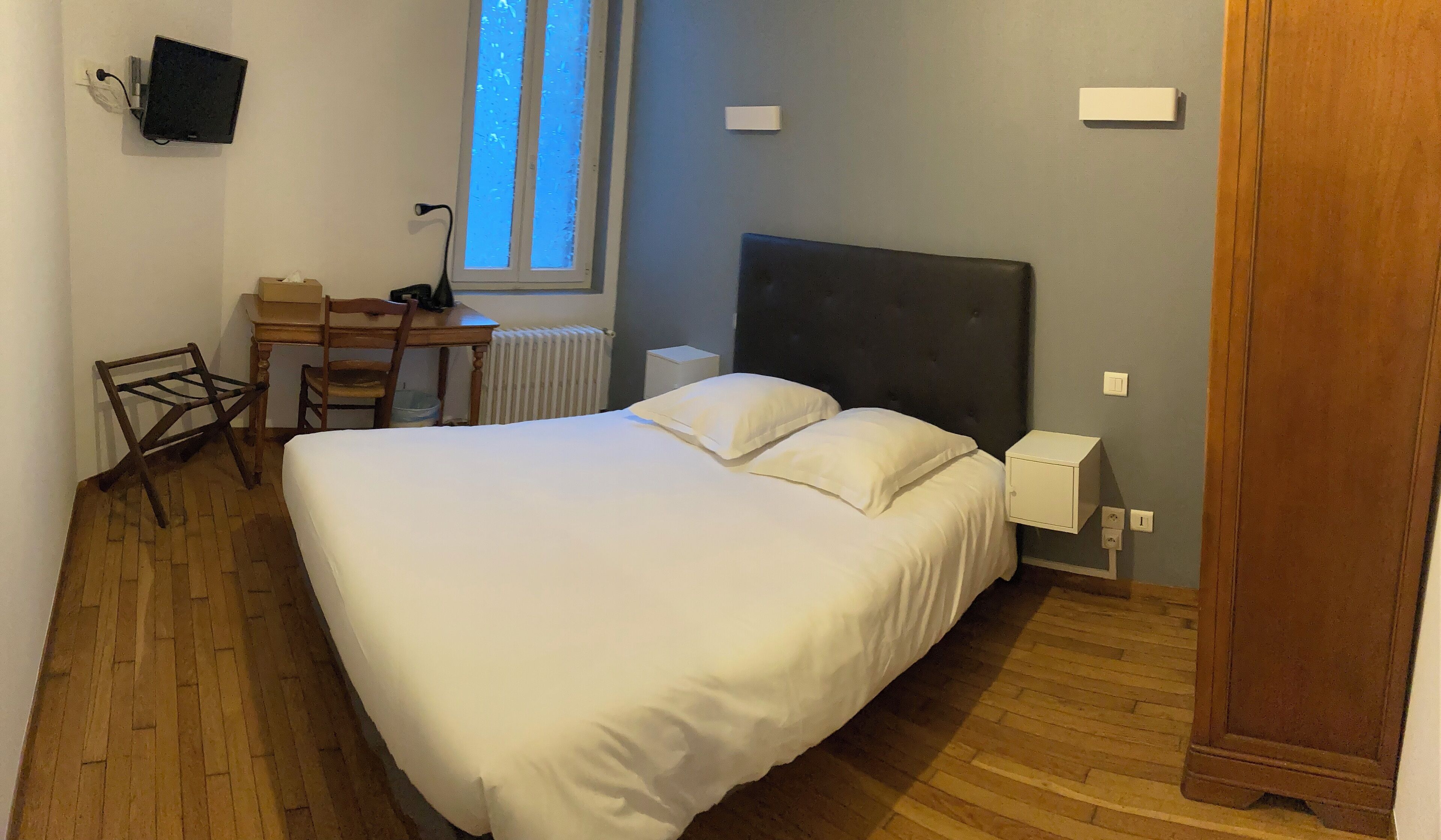 Une chambre à coucher avec un grand lit, un bureau, une chaise, un radiateur, une fenêtre et un téléviseur fixé au mur.