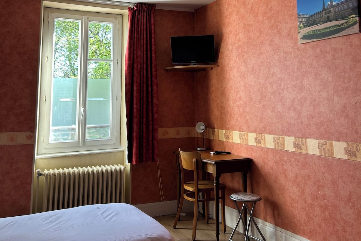 Quarto de hotel com duas camas, um ventilador de teto, uma TV fixada na parede, uma mesinha com uma cadeira e uma janela com cortinas.