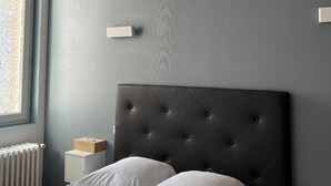 Une chambre à coucher avec une tête de lit capitonnée, du linge de lit blanc, une table de chevet et un radiateur.