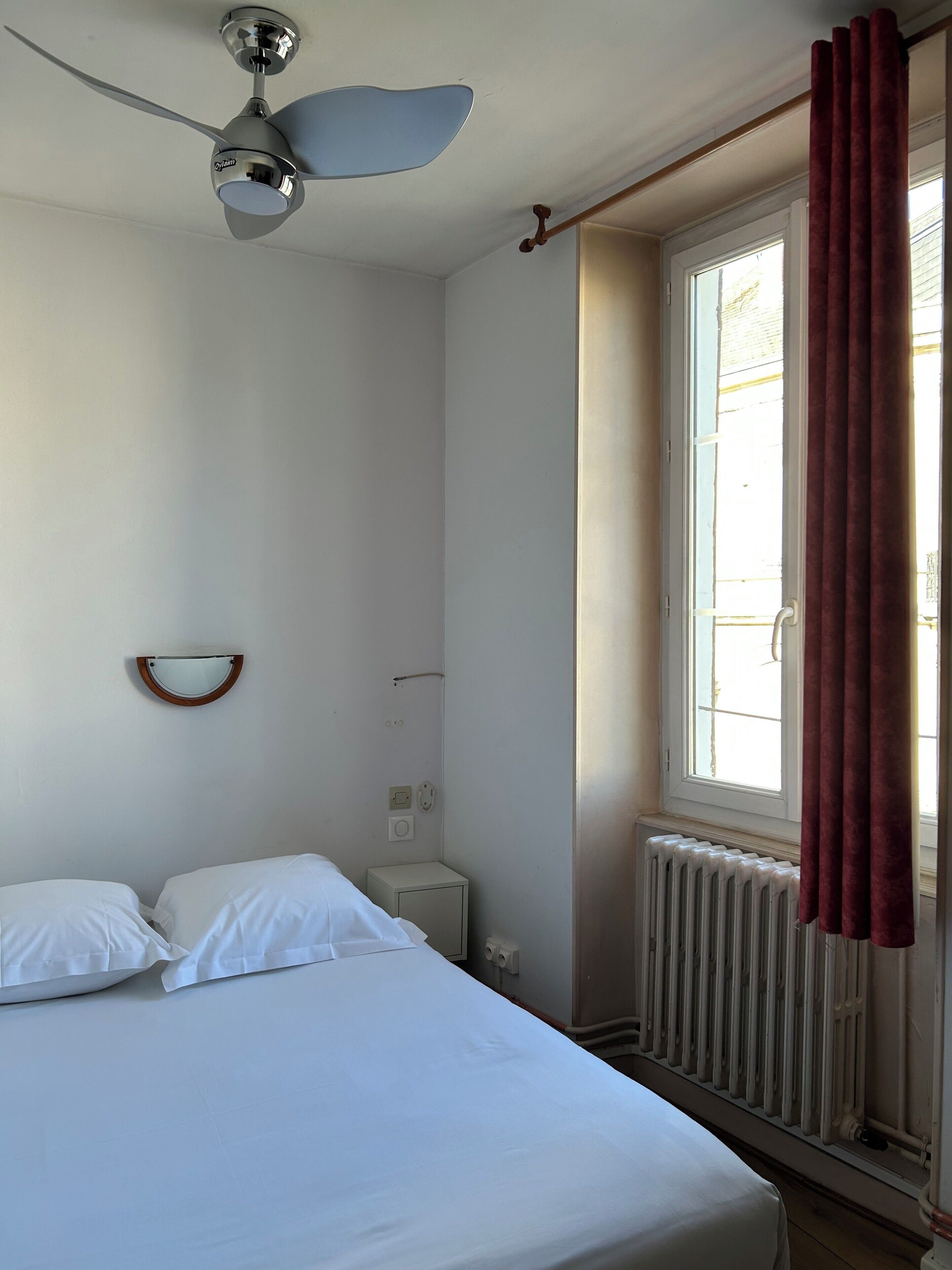 Un dormitorio con cama, ventilador de techo, ventana con cortinas, radiador y una mesita de noche pequeña.