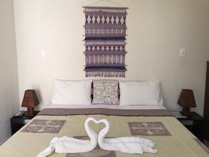 Cama bem arrumada com arranjo de toalhas em formato de coração, um pendurar de parede estampado e duas luminárias de cabeceira.