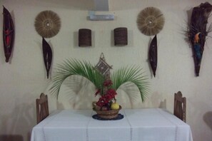 Uma área de refeições com uma mesa posta para dois, rodeada por cadeiras. Há penduras decorativas na parede e um pequeno arranjo de plantas sobre a mesa.