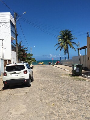 Uma rua pavimentada com uma SUV branca estacionada, uma palmeira e uma praia ao fundo.