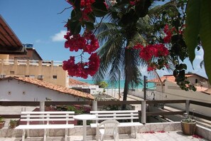 A terrace with white furniture, a palm tree, and a view of the beach and buildings. - Dolphin Pousada Gasthaus (Maragogi)