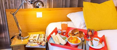 Una cama bien hecha con un almohadón amarillo, una mesita de noche con una lámpara, una bandeja con comida para el desayuno y una revista.