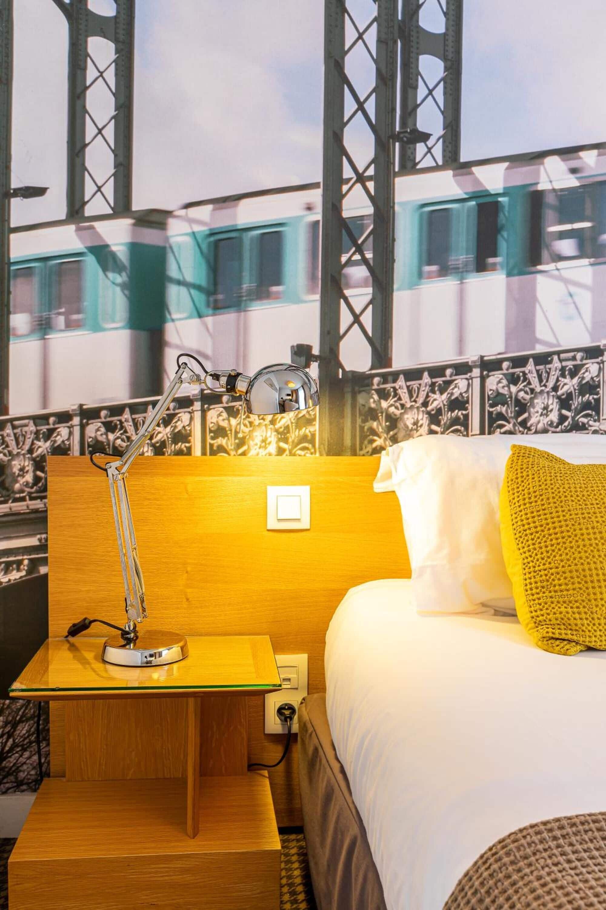 A hotel room with a bed, a nightstand, a lamp, and a view of a bridge and train.