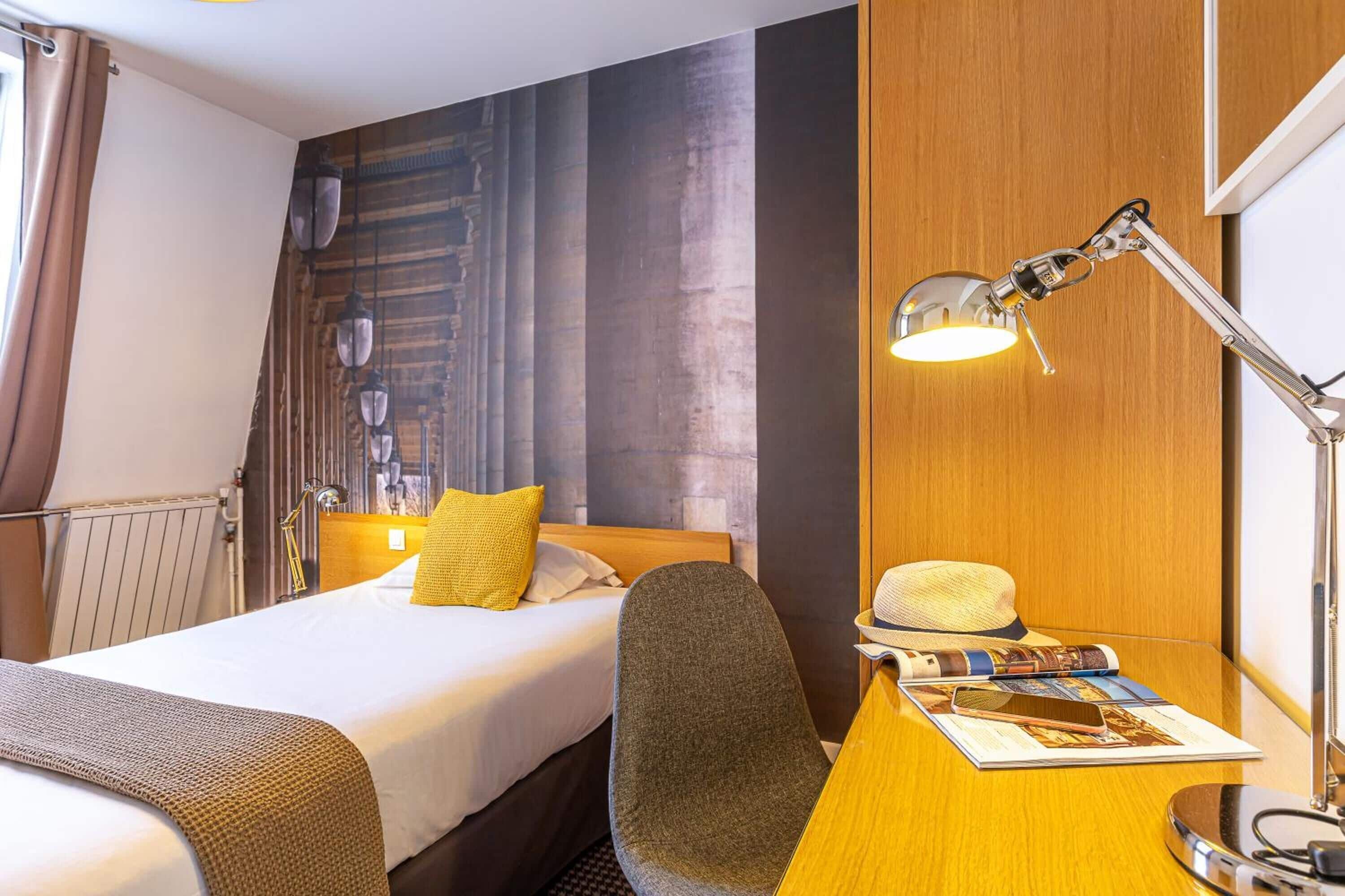 A hotel room with a bed, a desk with a lamp, a chair, and a hat on the desk.