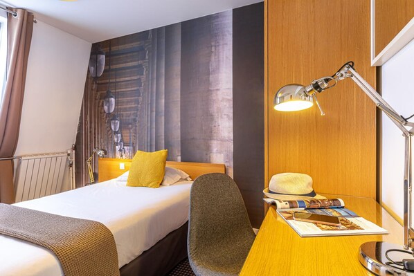 A hotel room with a bed, a desk with a lamp, a chair, and a hat on the desk. - Le 20 Prieure Hotel (Paris)