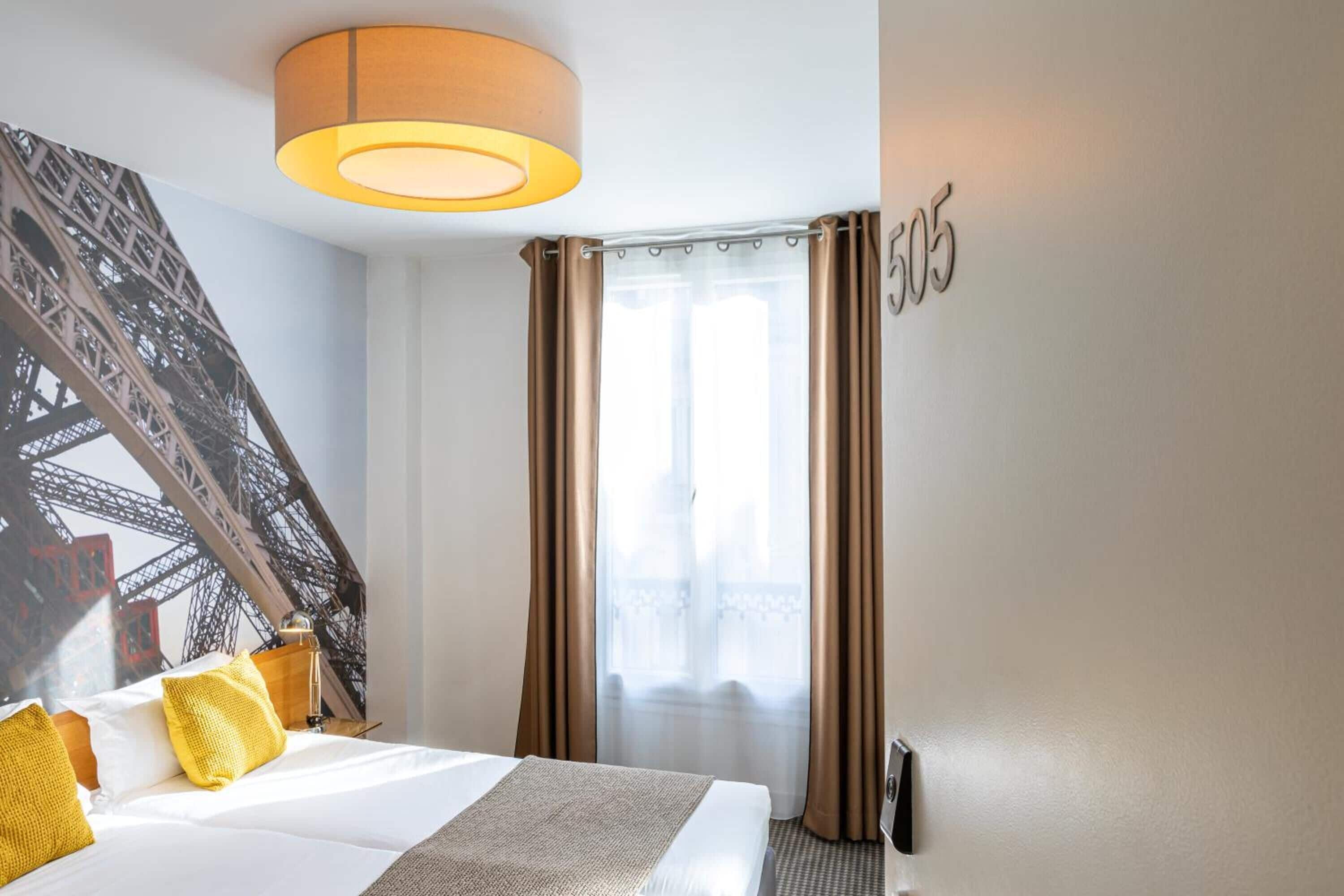 A hotel room with a bed, a lamp, a window with curtains, and a wall with a number.