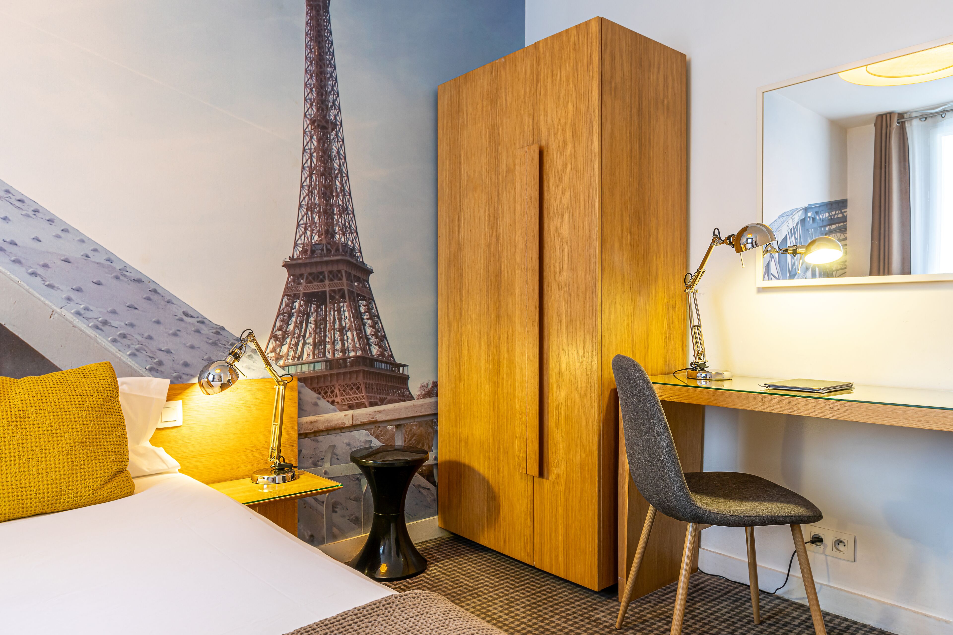 A hotel room with a bed, a desk with a chair, a lamp, a wardrobe, and a mural of the Eiffel Tower.