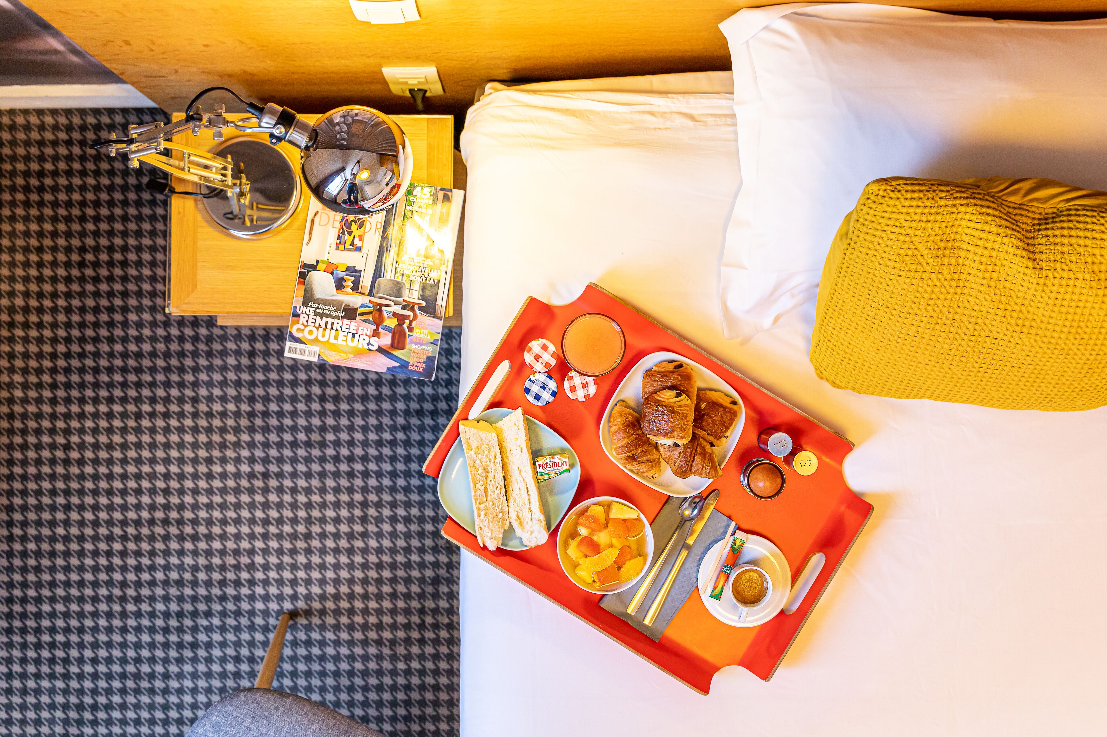 Quarto de hotel com cama, bandeja de café da manhã, uma revista e uma mesinha com abajur.