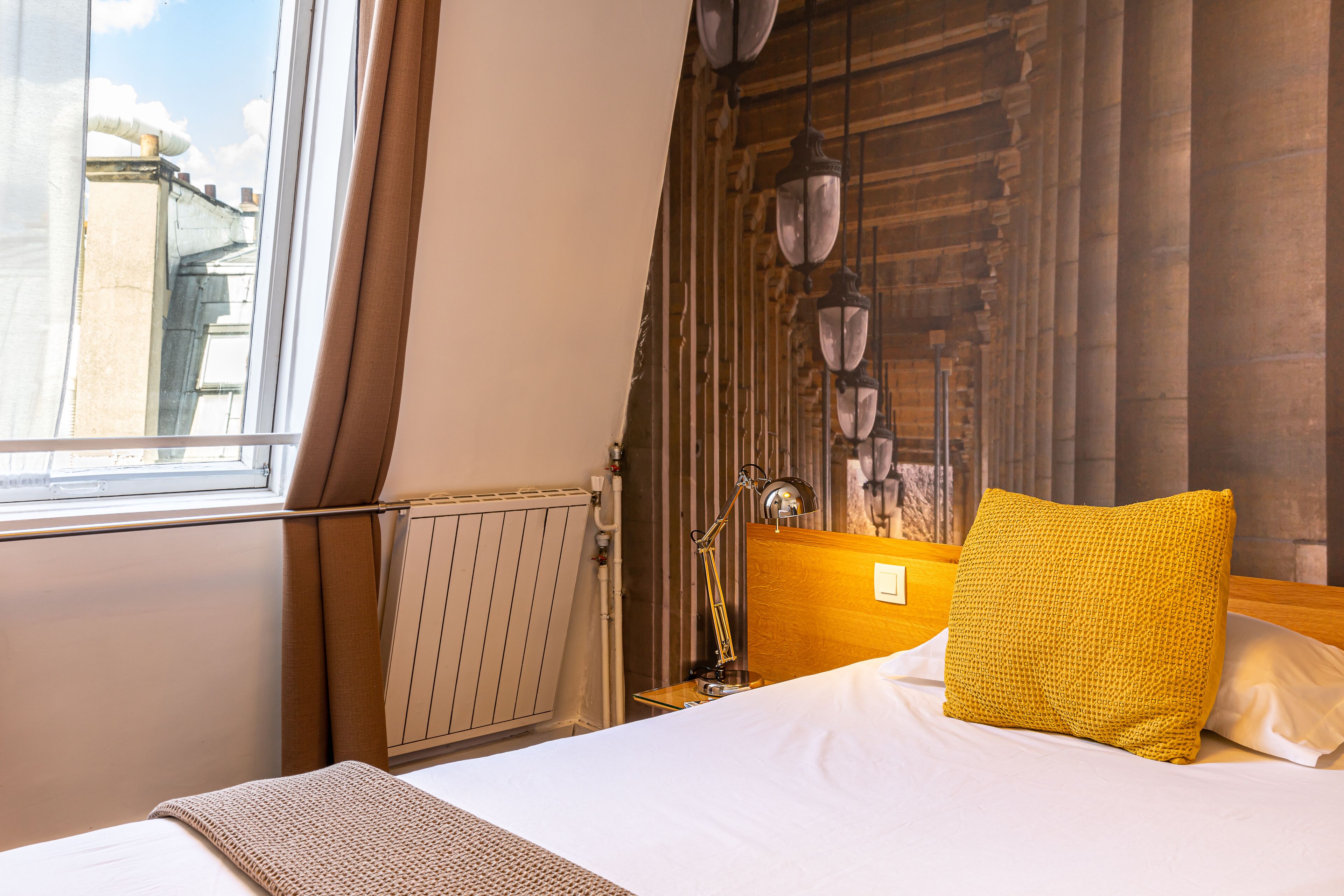 Una habitación de hotel con cama, ventana con cortinas, radiador y pared con lámparas colgantes.