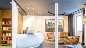 Una moderna zona de recepción con un mostrador curvo, un espacio de espera con un sillón de cuero marrón, una estantería con diversos objetos y un cuadro de un paisaje urbano colorido en la pared.
