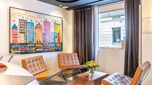 Una habitación de hotel moderna con un cuadro grande de un paisaje urbano colorido, dos sillones de cuero, una mesa de centro de vidrio con un libro y vista a un edificio a través de una ventana.