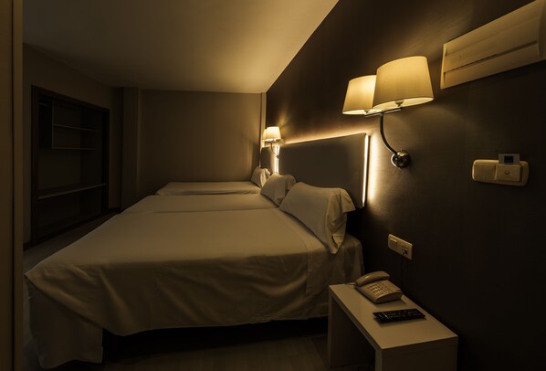 A hotel room with two beds, a bedside table with a phone and remote control, and wall-mounted lamps.