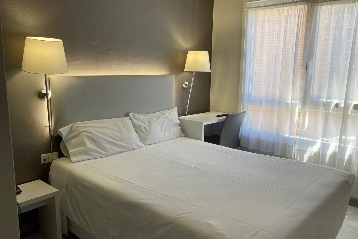 A hotel room with a large bed, two bedside lamps, a small table, and a window with sheer curtains.