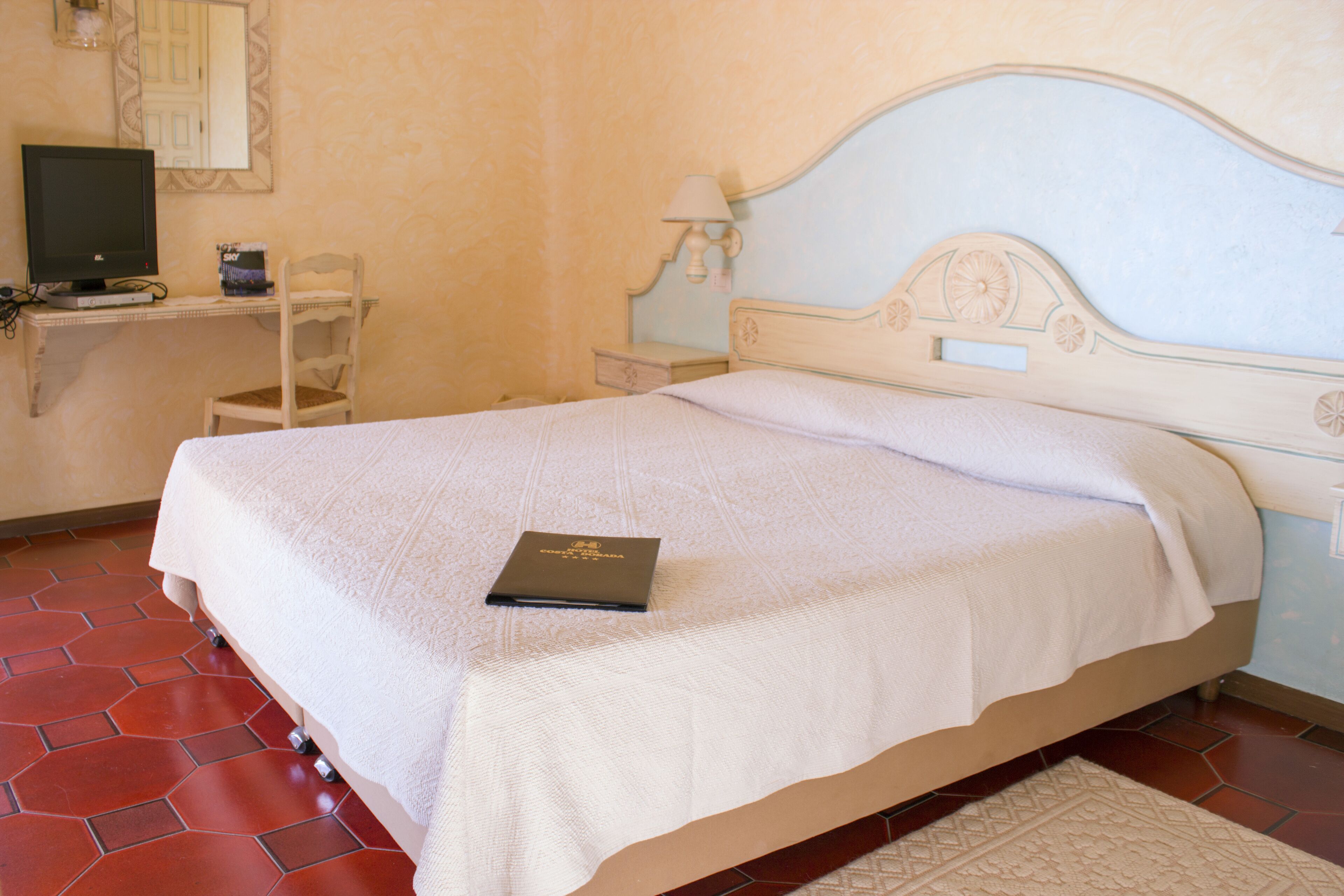 Un dormitorio con una cama, un escritorio con computadora, una silla y una mesita de noche con lámpara.