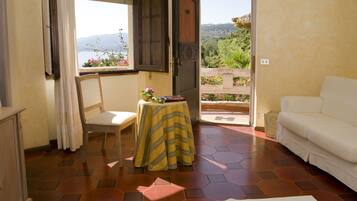 Una habitación con un sofá blanco, una mesa pequeña con mantel amarillo, una silla y vistas a un jardín a través de una puerta abierta.