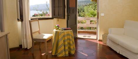 A room with a white sofa, a small table with a yellow tablecloth, a chair, and a view of a garden through an open door.