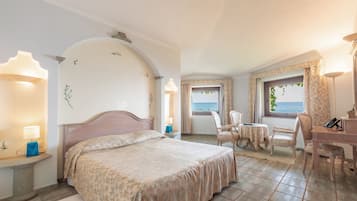 A bedroom with a bed, bedside tables, a desk, and chairs. There is a view of the sea through the window.