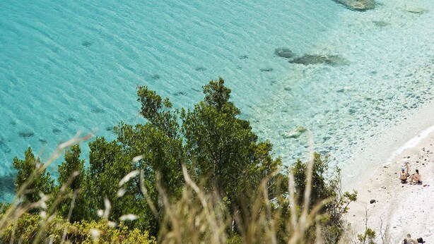 A boat on clear turquoise water near a rocky shoreline with a beach in the foreground. - Hotel Costa Dorada (Dorgali)
