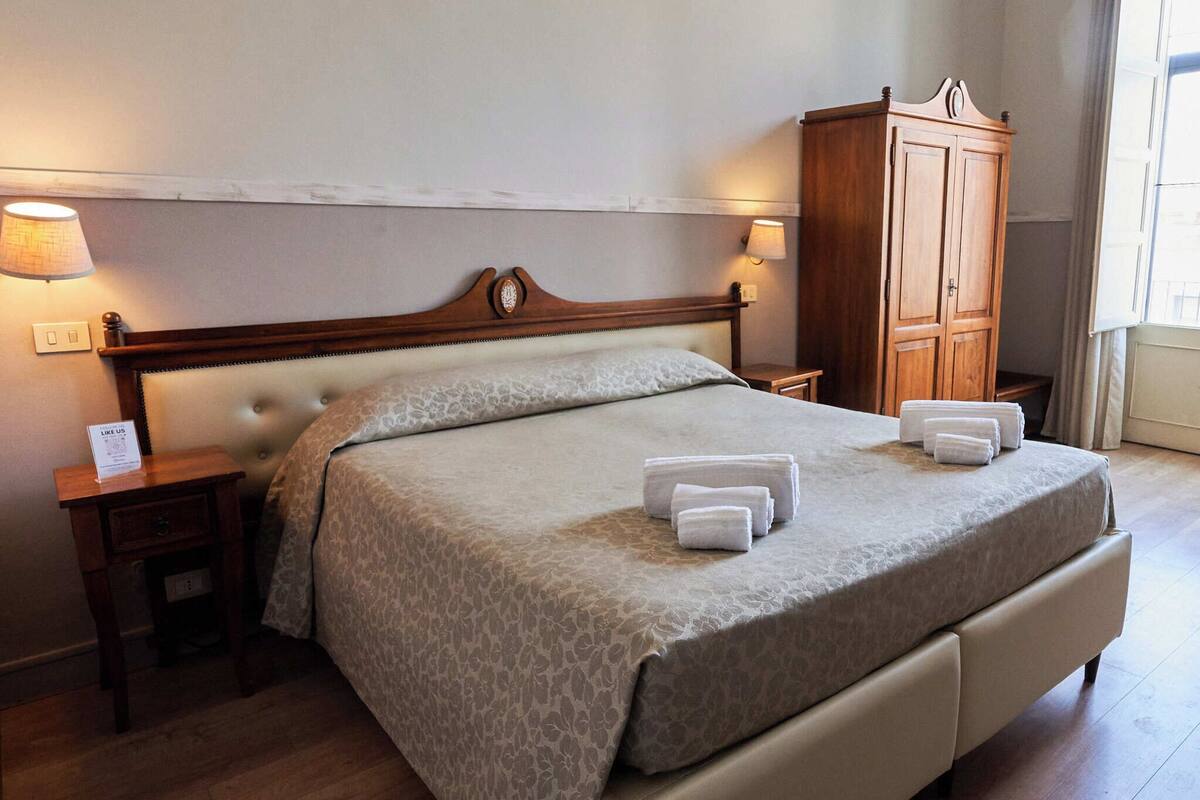 A neatly made bed with a headboard, two bedside tables with lamps, a wooden wardrobe, and a window with curtains.