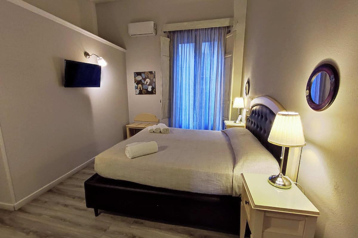 A hotel room with a bed, bedside table, lamp, air conditioning unit, TV, and a window with curtains.