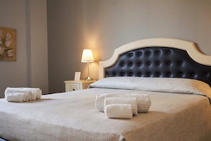 Una cama bien hecha con un cabecero capitonado, una lámpara de noche y toallas enrolladas sobre la cama.