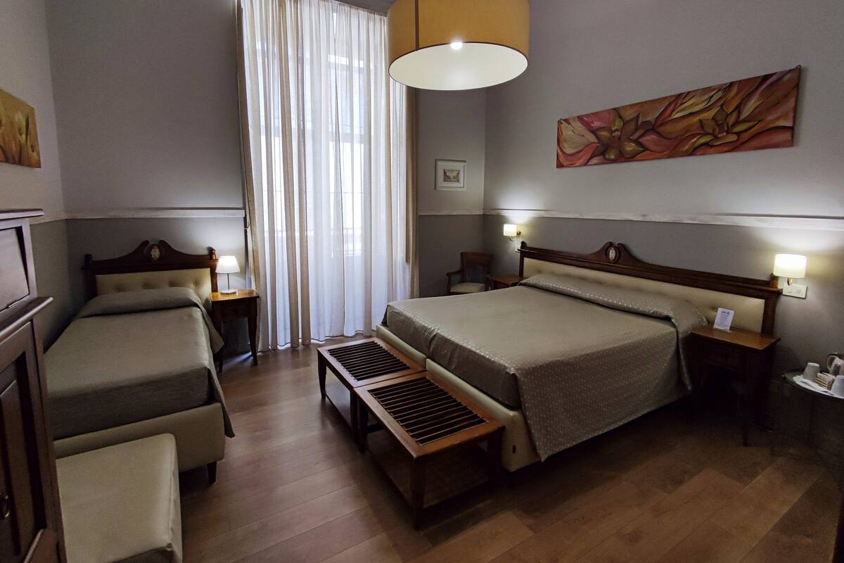A hotel room with a large bed, two smaller beds, a desk, a chair, a window with curtains, and a painting on the wall.