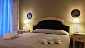 A neatly made bed with rolled towels, two bedside tables with lamps, a round mirror, and a window with blue curtains. - La Collegiata Guest House (Catania)