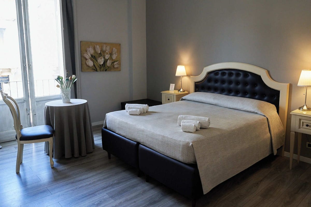 A neatly arranged hotel room with a large bed, two bedside tables with lamps, a chair, and a small table with a vase of flowers.