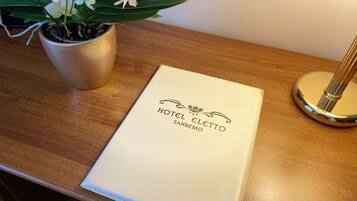 Quarto de hotel com uma escrivaninha de madeira, uma orquídea em vaso e um menu intitulado "HOTEL ELETTO SANREMO".