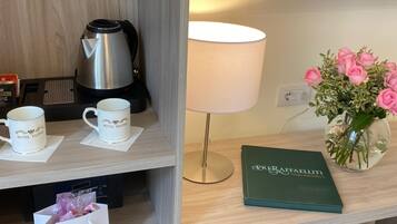 Une chambre d’hôtel avec un bureau en bois, une machine à café, deux tasses, une lampe, un livre, un vase avec des fleurs et une petite table sur laquelle se trouve un sac.