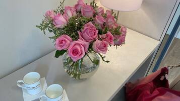 Una mesa blanca con un jarrón de rosas rosadas, dos tazas blancas con detalles dorados y un bolso rojo.