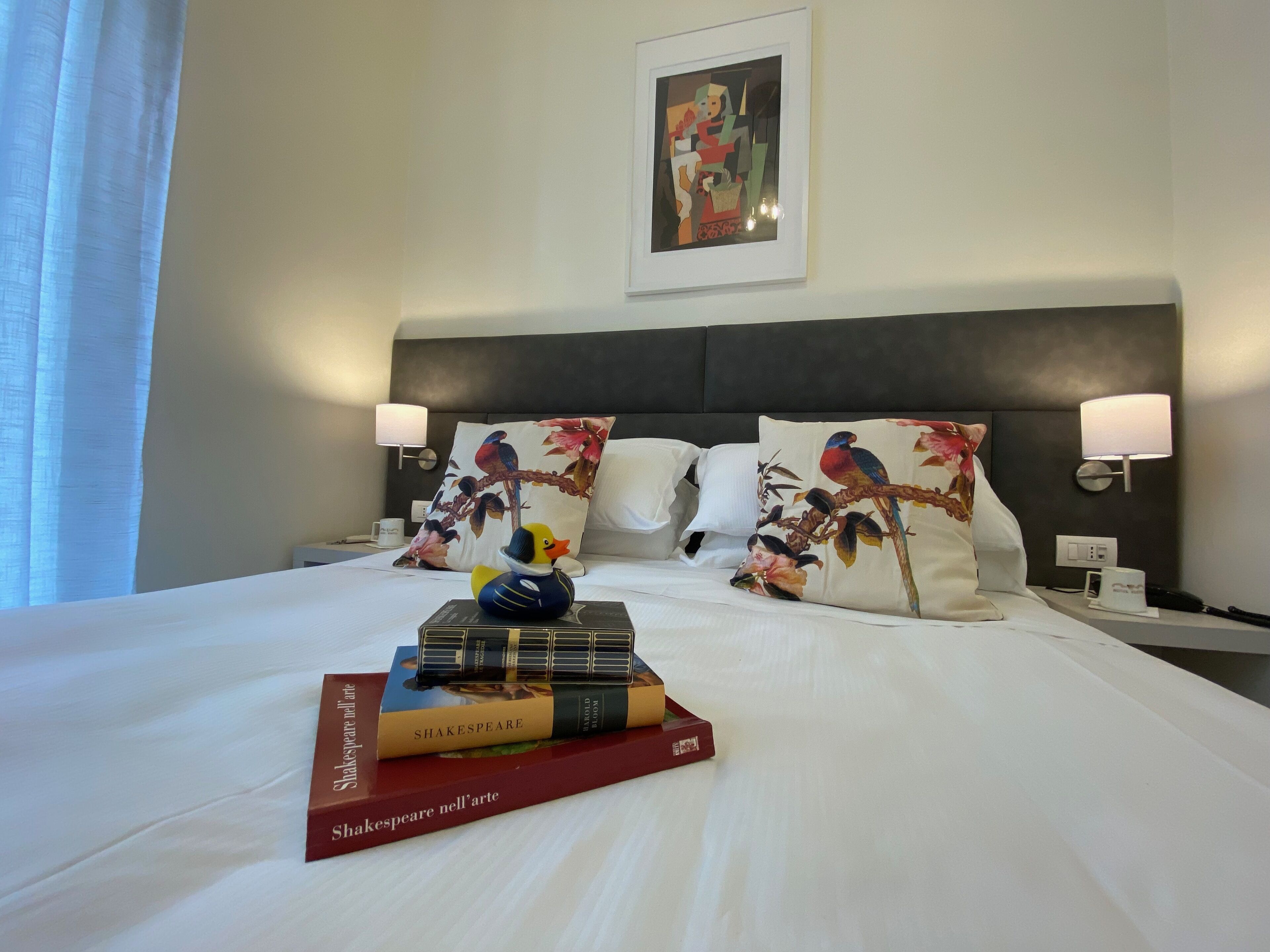 Un lit bien fait, avec des oreillers, un coussin décoratif orné d’un motif d’oiseau, une petite figurine de canard et une pile de livres.