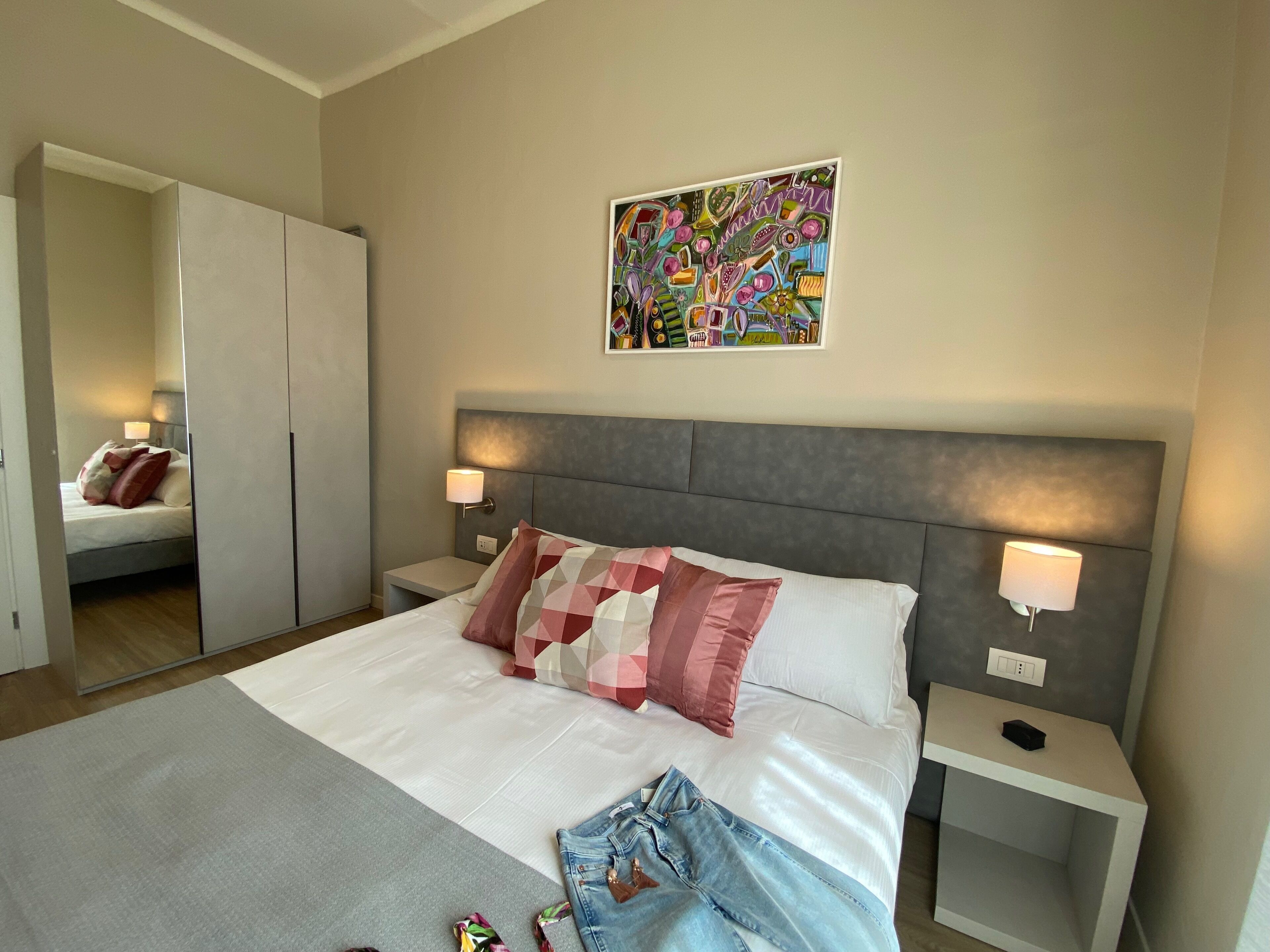 A hotel room with a bed, two bedside tables, a wardrobe, and a colorful abstract painting on the wall.