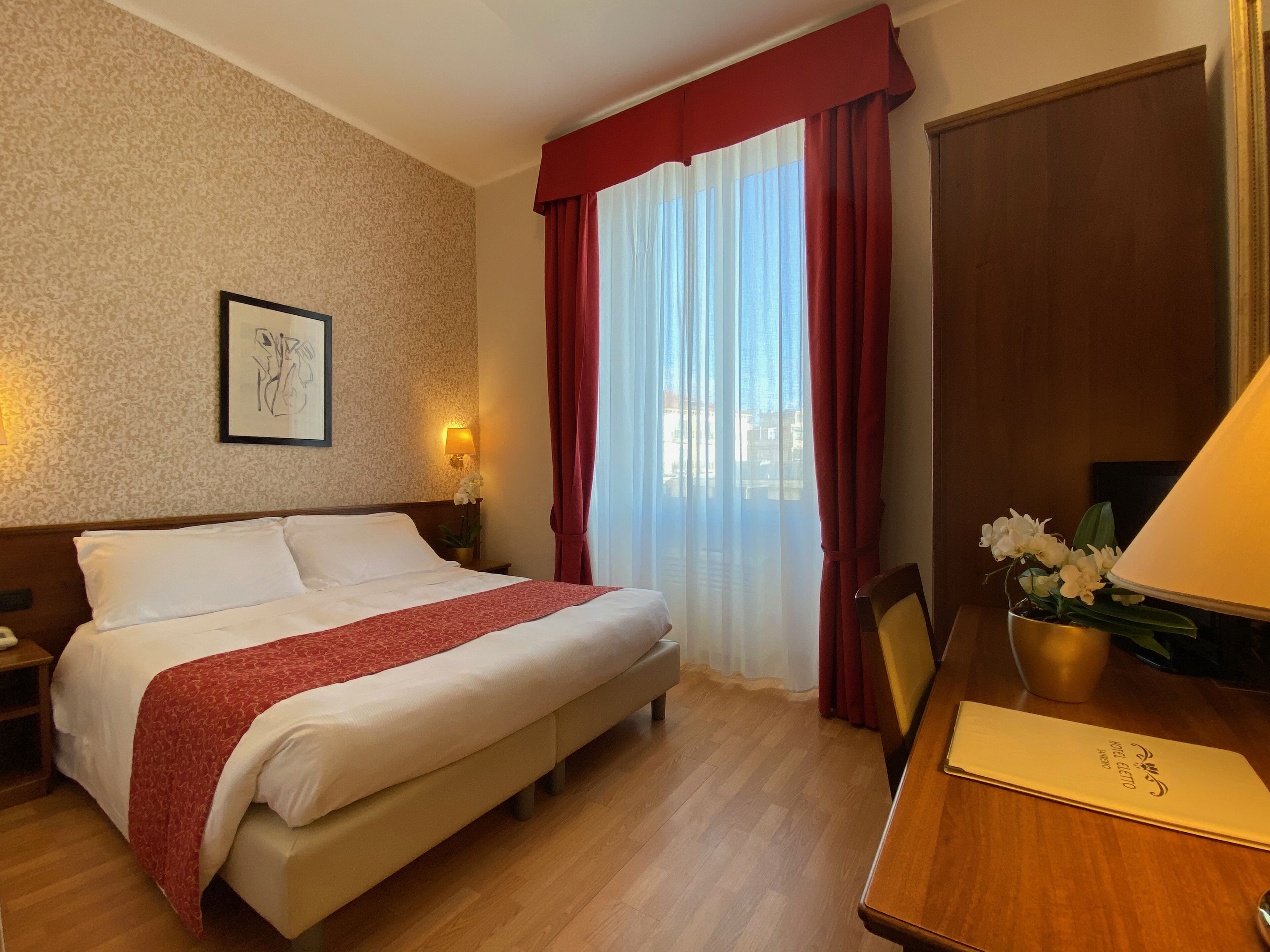 A hotel room with a bed, a desk, a lamp, a framed picture on the wall, and a window with curtains.