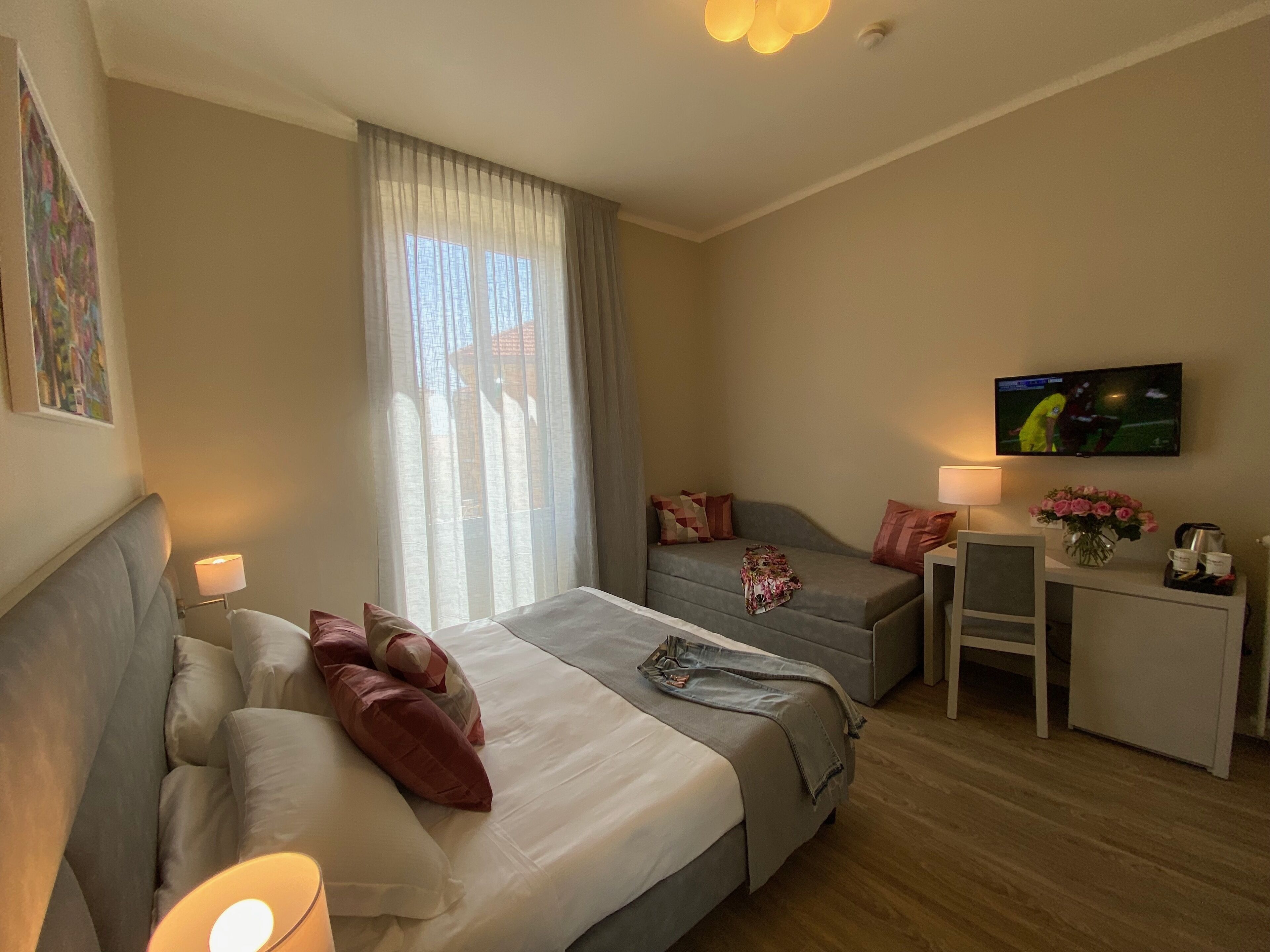 A hotel room with two beds, a sofa, a TV, a desk with a lamp, and a vase of flowers.