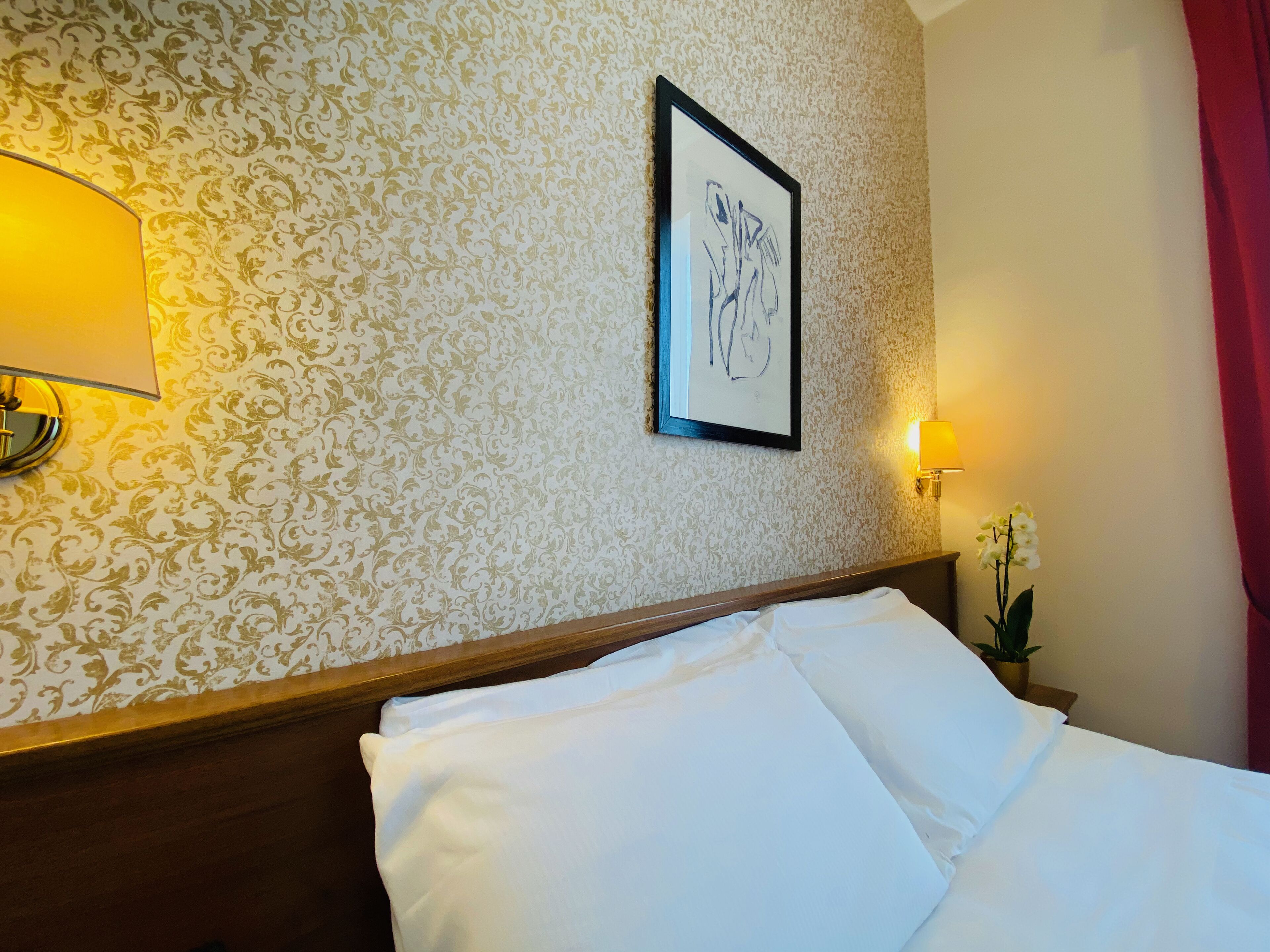 Una habitación de hotel con una cama, una pared con papel tapiz estampado, una obra de arte enmarcada y una mesita de noche con una lámpara y una planta en maceta.