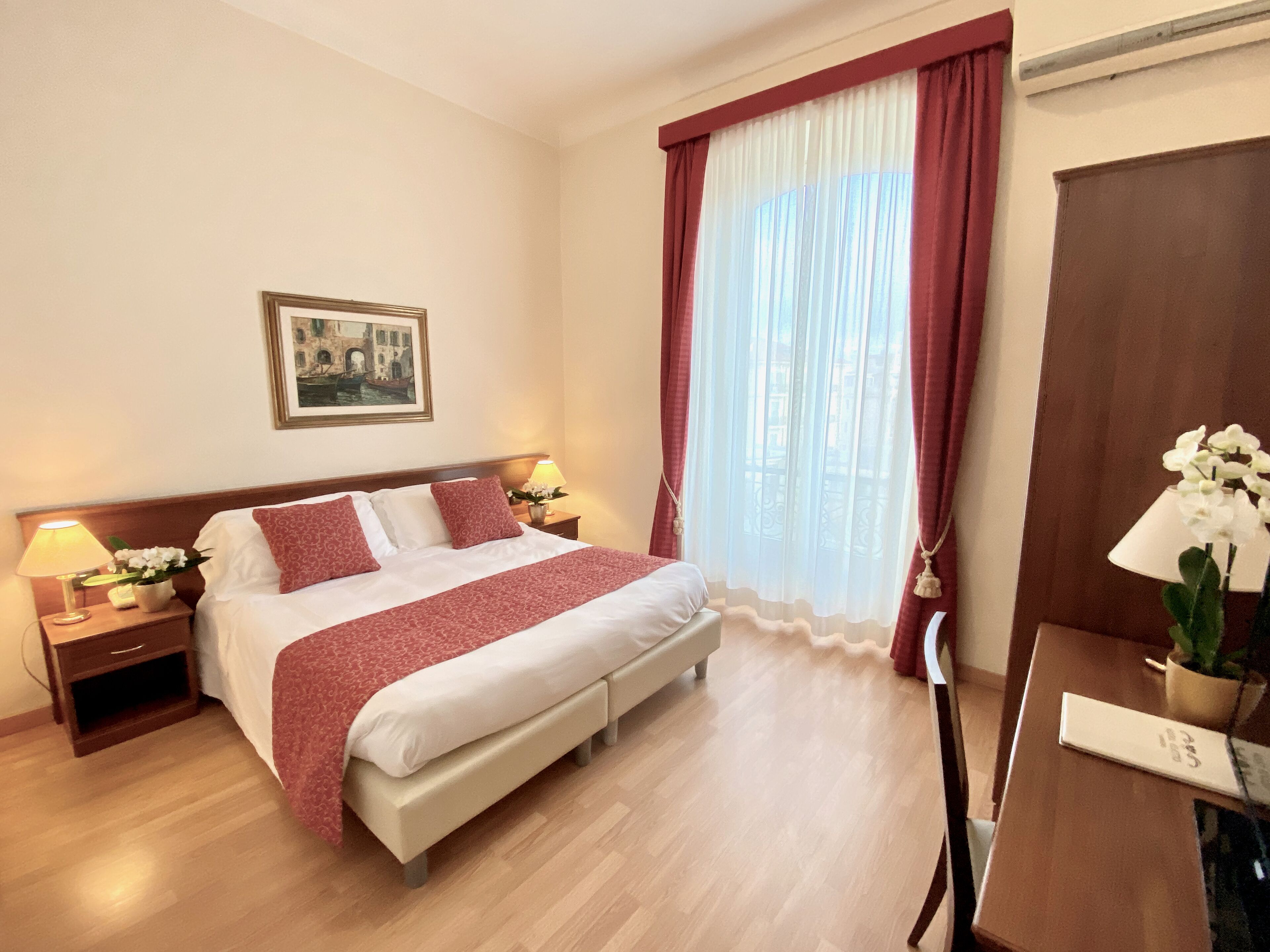 Een hotelkamer met een groot bed, twee nachtkastjes met lampen, een bureau met stoel, een raam met gordijnen en een ingelijst schilderij aan de muur.