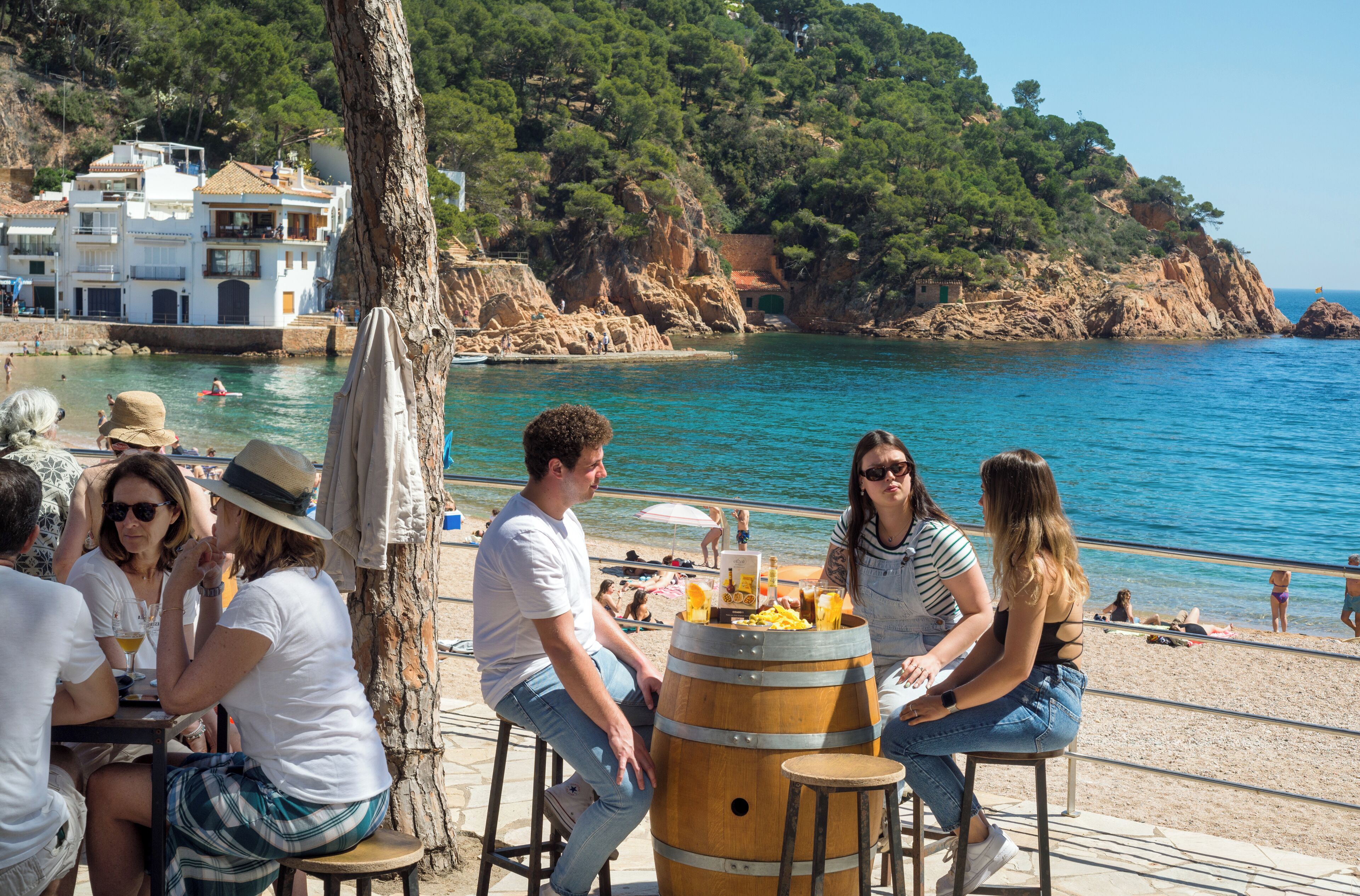 Un grupo de personas sentadas en una mesa al aire libre junto a la costa, con vista al mar y una colina rocosa al fondo.