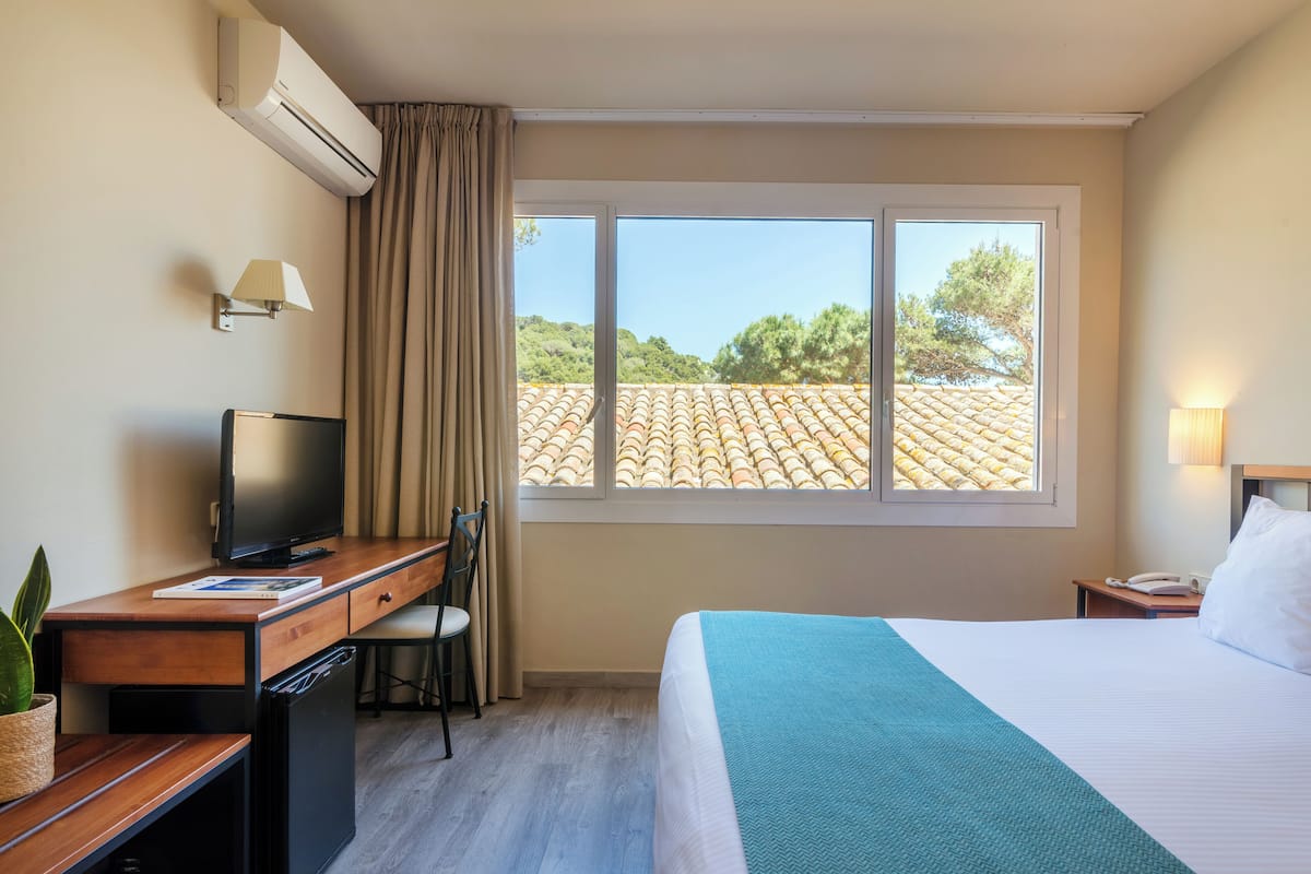 A hotel room with a bed, a desk with a computer, a chair, a TV, a window with curtains, and a lamp.