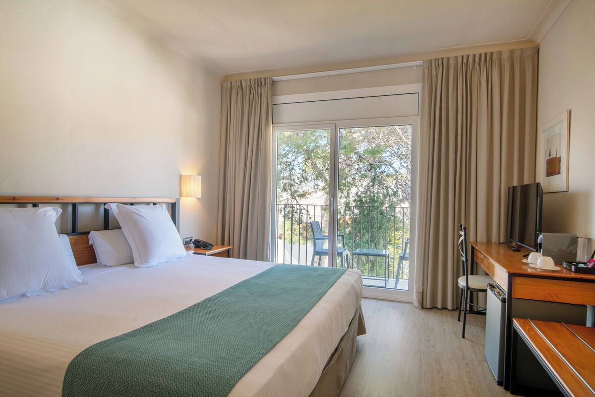 A hotel room with a bed, a desk, a chair, a TV, and a balcony with curtains.