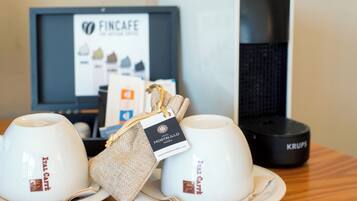 Deux tasses blanches portant l’inscription « ITAL CAFE » sont posées sur une table en bois. On aperçoit également un objet enveloppé de toile de jute avec une étiquette d’hôtel, ainsi qu’une machine à café Krups en arrière-plan.
