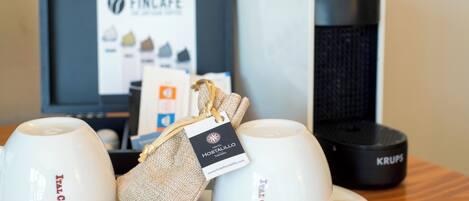 Two white cups with "ITAL CAFE" labels on a wooden table, a burlap-wrapped item with a hotel tag, and a Krups coffee maker in the background.