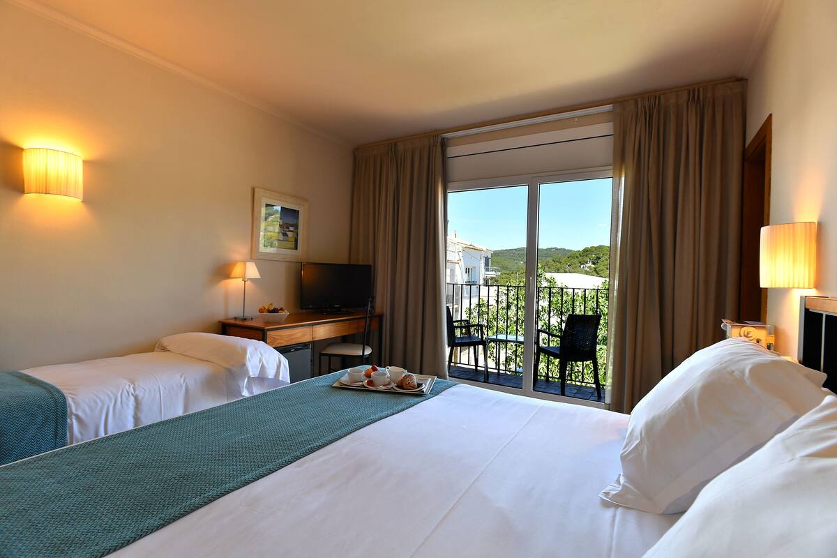 A hotel room with two beds, a desk with a television, and a balcony with a view of greenery.