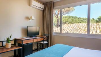 Une chambre d’hôtel comprenant un lit, un bureau avec une télévision, une chaise, un petit réfrigérateur, une plante en pot et une fenêtre donnant sur un toit recouvert de tuiles et sur de la verdure.